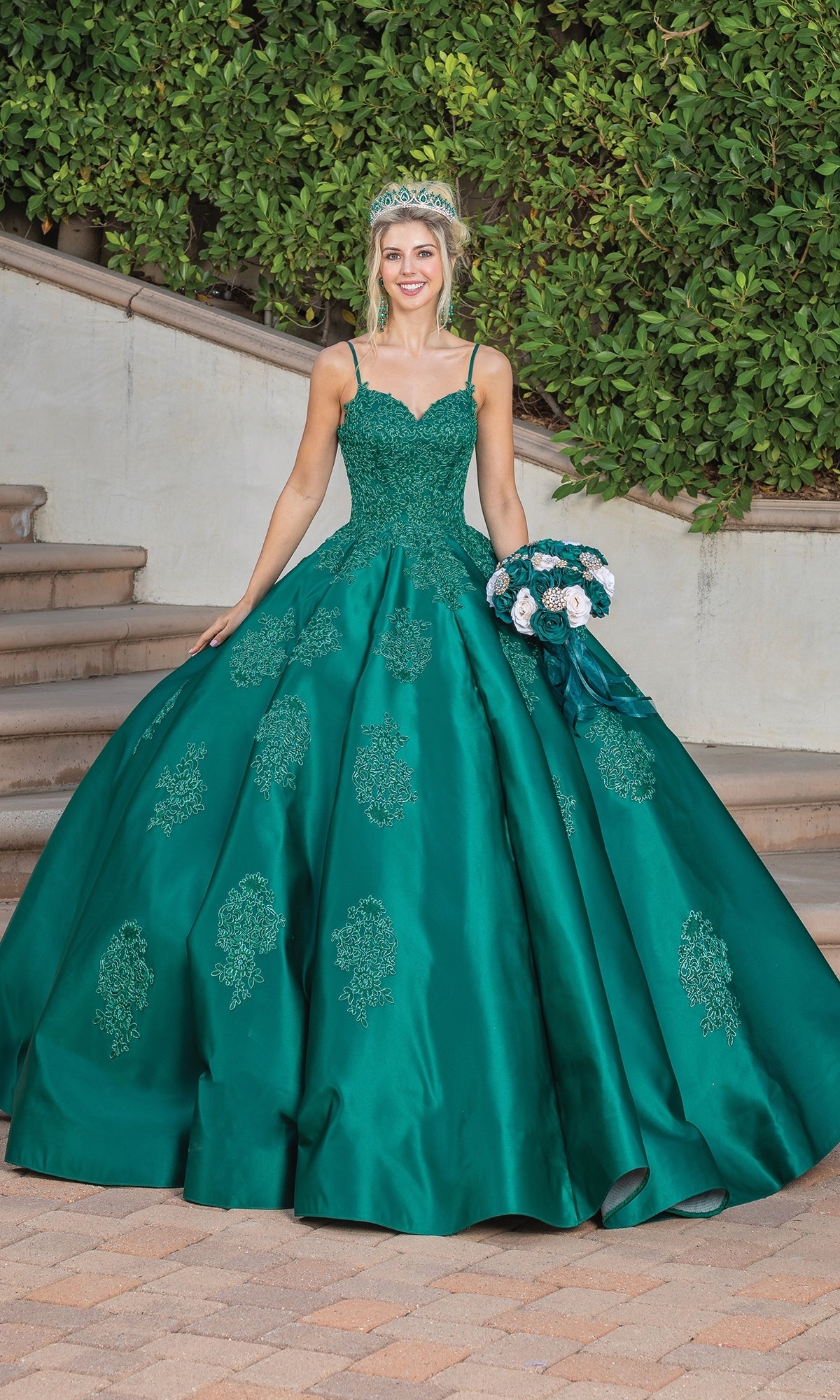 A young woman stands on a stone path outdoors, holding a bouquet of white and teal flowers while wearing the Quinceanera Dress 1325 by Dancing Queen, an a-line ball gown with a sweetheart neckline and lace applique.